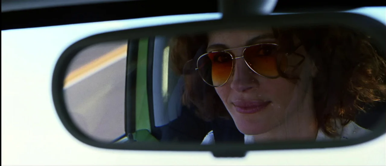 The Mexican (2001) movie still: closeup — The interior of a moving vehicle features a driver viewed through a rearview mirror. The reflection shows…