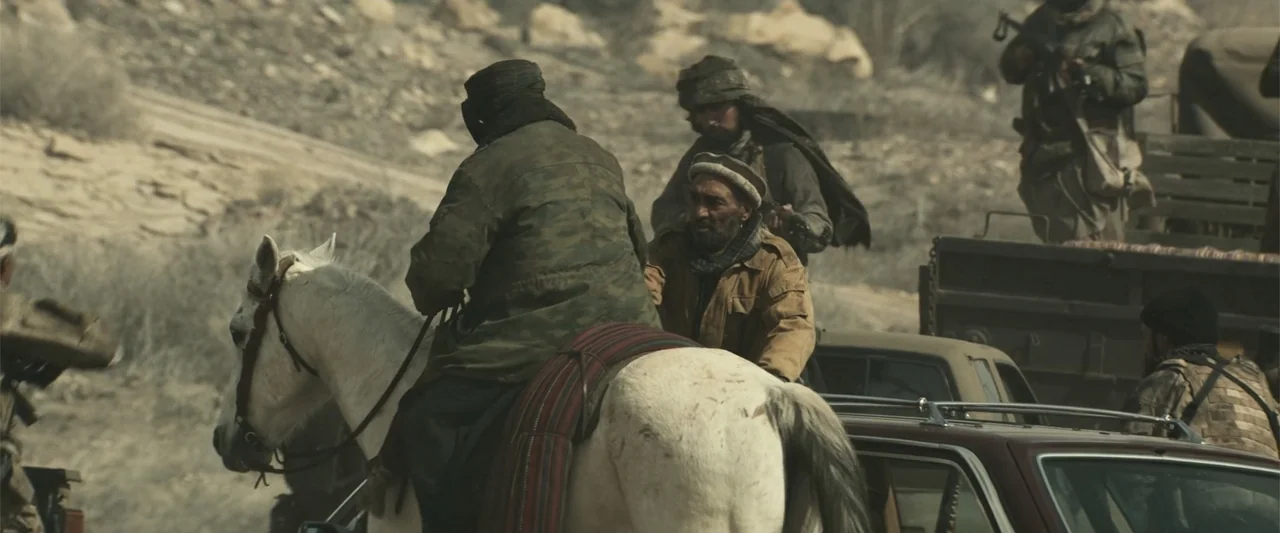 12 Strong (2018) movie still: wide — An arid rocky landscape contains several armed men on horseback and in pickup trucks. The group travels across…