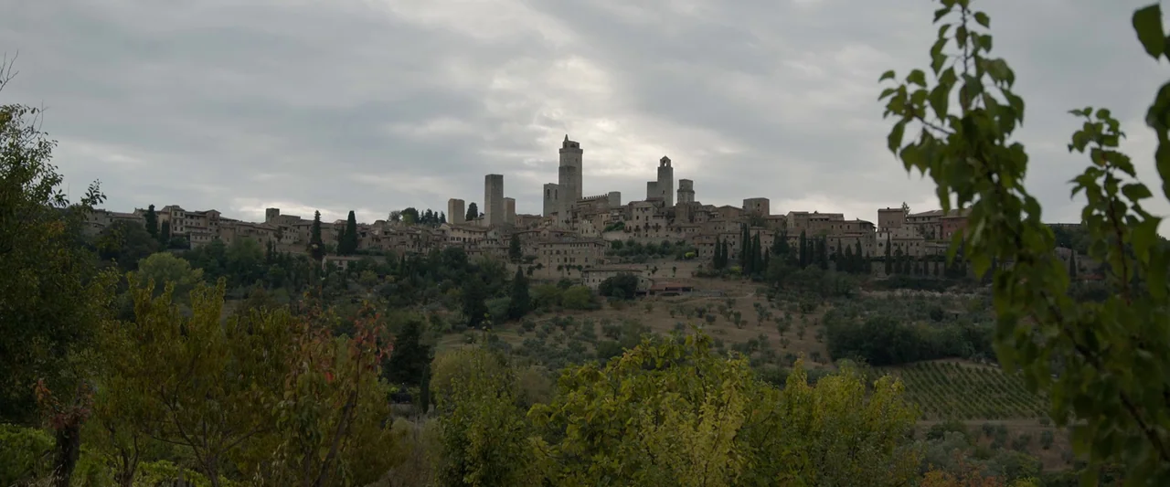 Rosaline (2022) movie still: establishing — A medieval walled town with tall stone towers sits atop a grassy Tuscan hill. Dense foliage, olive…