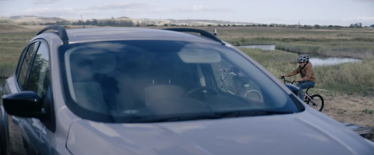 A Christmas Mystery (2022) movie still: wide — A silver car occupies the foreground with blurred marshland and rolling hills behind. Two children in…