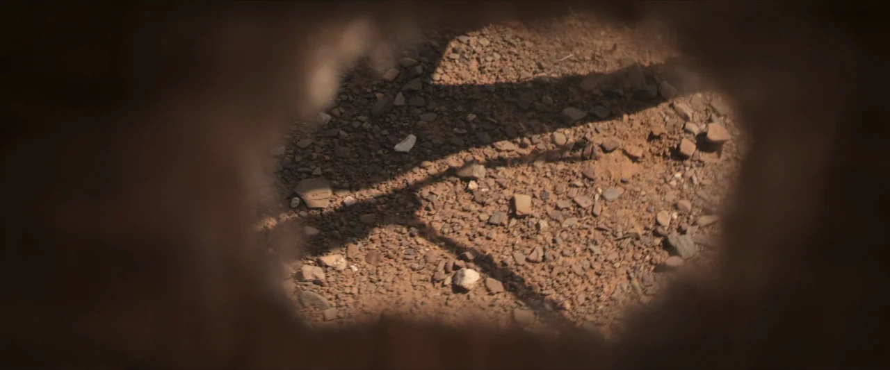 The New Boy (2023) movie still: closeup — The ground consists of sun-baked red dirt covered in scattered small rocks and pebbles. A long, thin…