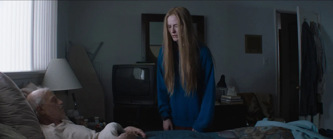 Kajillionaire (2020) movie still: medium — A dimly lit room contains an elderly man lying in a bed with medical equipment. A young woman with long…