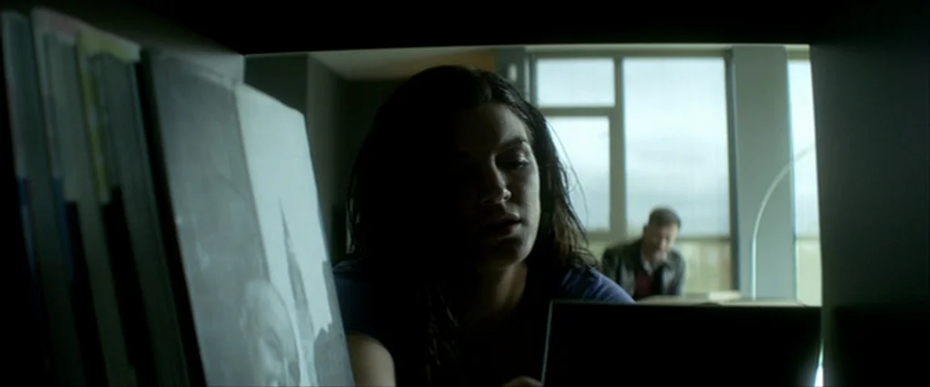 Haywire (2011) movie still: medium — A modern room features a shelf in the foreground containing several books or records. A woman sits in the…