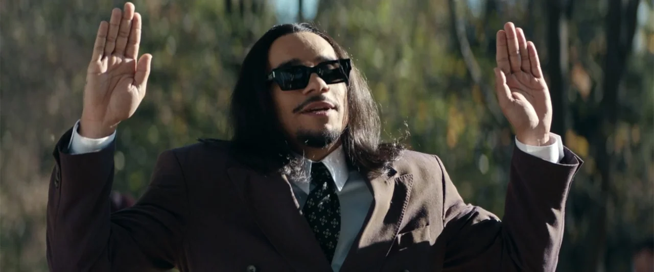 McWalter (2025) movie still: medium — An outdoor area with blurred trees and foliage providing a natural backdrop. A man in a brown suit holds both…