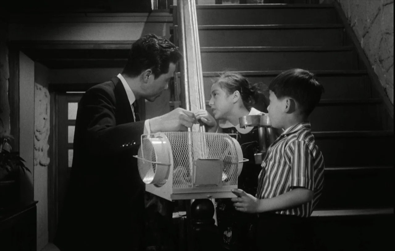 The Housemaid (1960) movie still: medium — A man and two children stand on a wooden staircase at night. They examine a small wire birdcage held by…