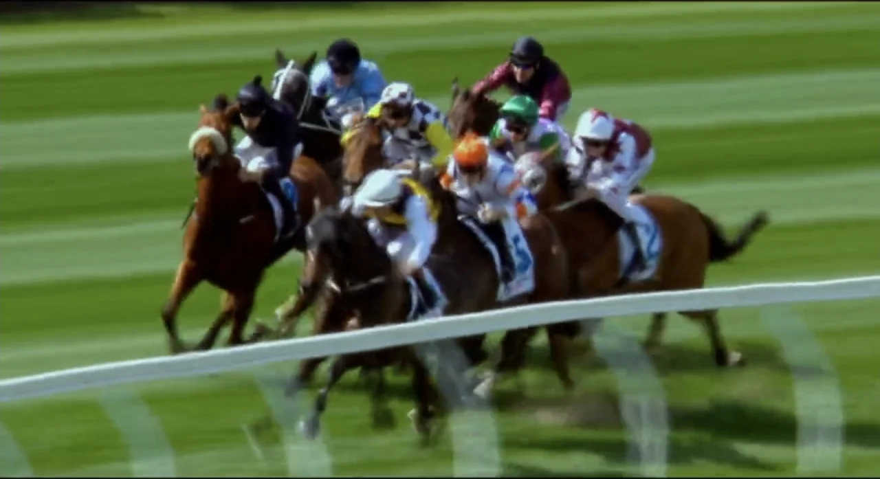 Ride Like a Girl (2019) movie still: wide — Multiple jockeys race horses across a green turf track under bright daylight. The group of riders leans…