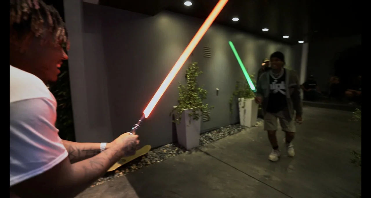 Juice WRLD: Into the Abyss (2021) movie still: medium — A modern outdoor patio with gray walls and potted plants at night. Two men engage in a light…