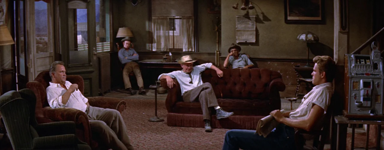 Bad Day At Black Rock (1955) movie still: wide — An interior lobby features dark velvet furniture and a vintage floor-standing slot machine. Four…