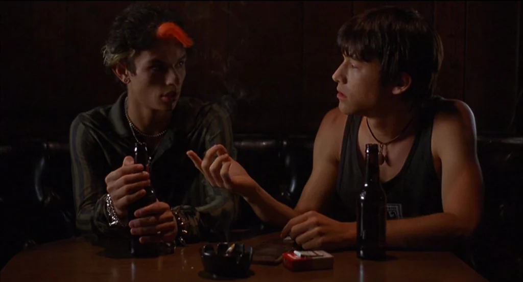 Mysterious Skin (2004) movie still: medium — A dimly lit booth with dark wood paneling in a bar or tavern. Two young men sit opposite each other…