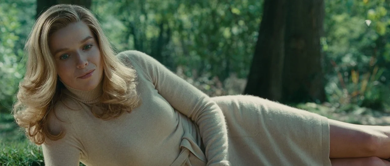 The American (2010) movie still: medium — A woman rests on the grass in a wooded area with dense greenery. She lies on her side, supported by one…