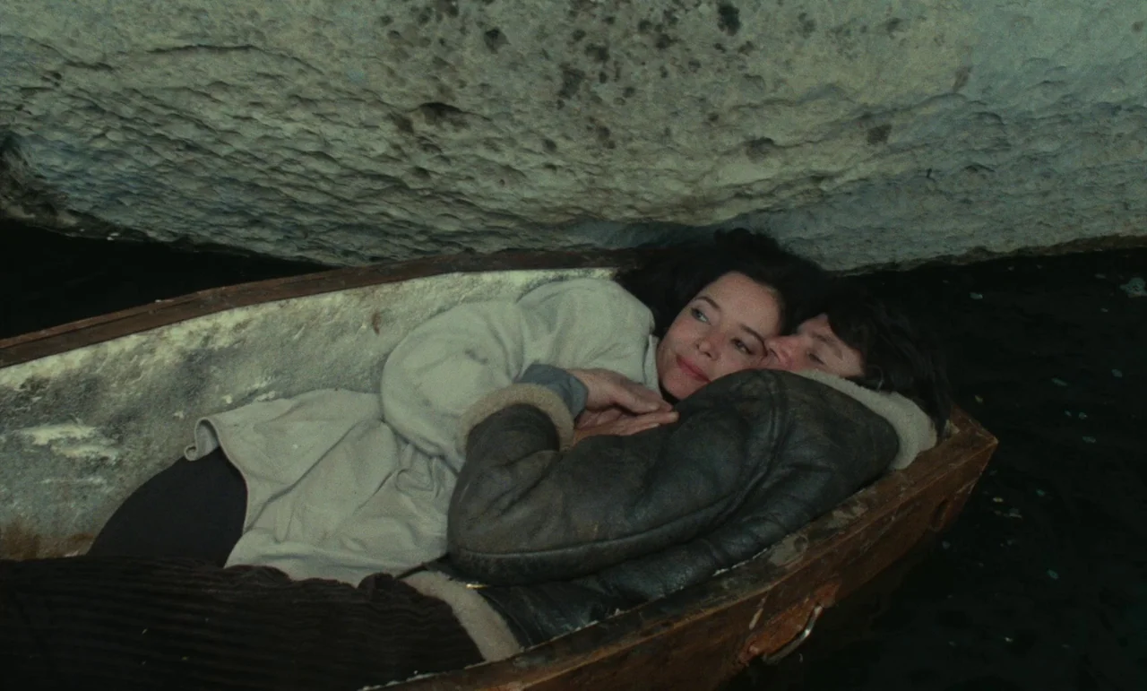 Litan (1982) movie still: medium — A man and a woman lie together in a cramped wooden boat inside a limestone cave. The low rocky ceiling hangs…