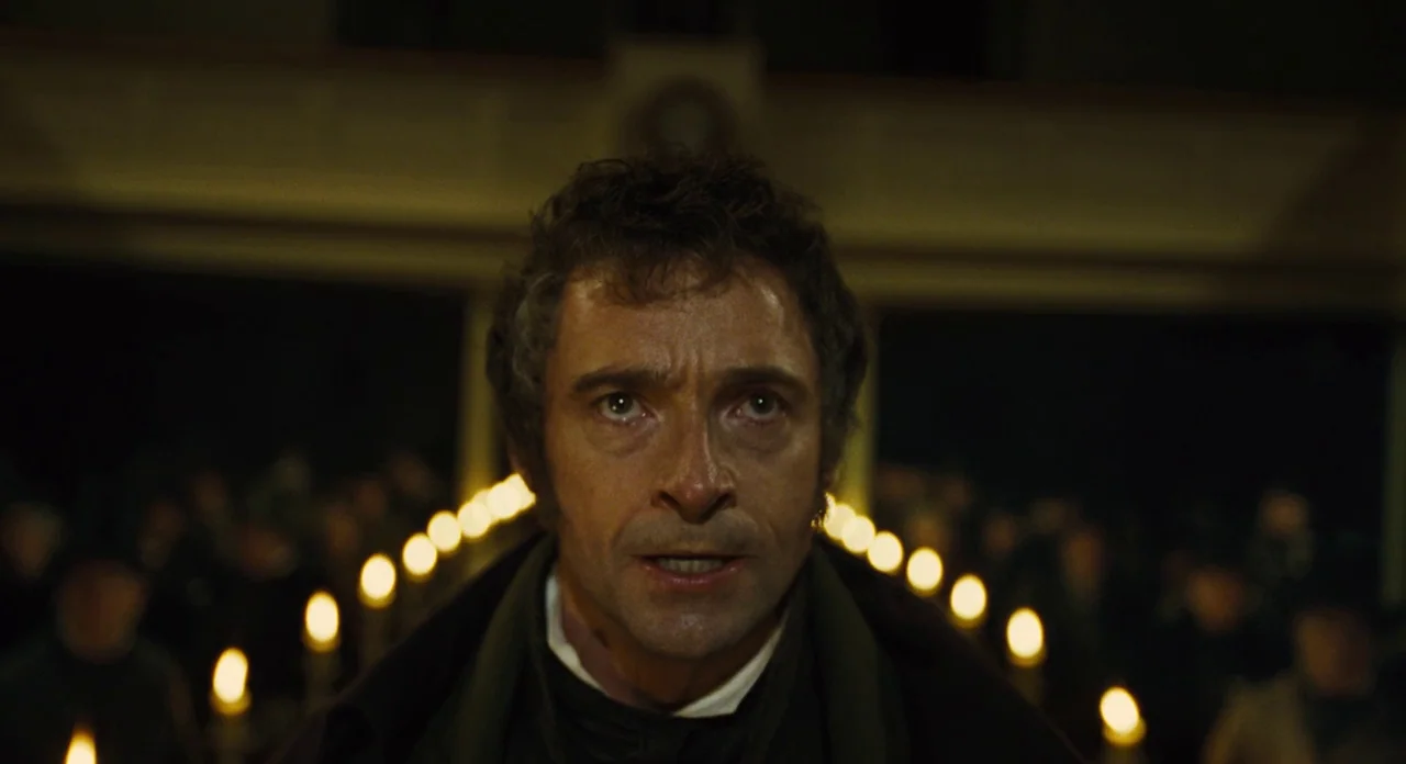 Les Misérables (2012) movie still: closeup — A dimly lit, expansive interior space with numerous small candle flames glowing in the background. A…