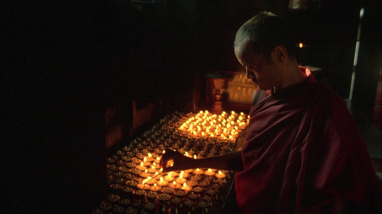 Powaqqatsi (1988) movie still: medium — A dimly lit room contains rows of small metal containers filled with burning candles. A man in a maroon robe…