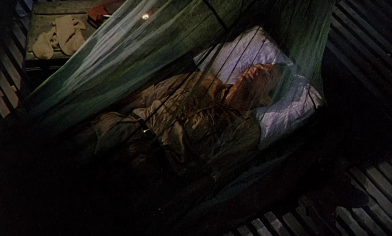 The Purple Plain (1954) movie still: medium — A man lies on a cot inside a dark, confined wooden structure at night. He is covered by a hanging…