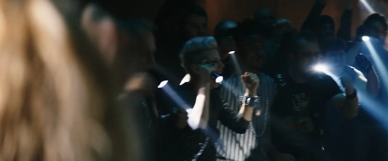 Jolt (2021) movie still: medium — A dark indoor space contains a group of people facing the camera. Several individuals hold bright handheld…