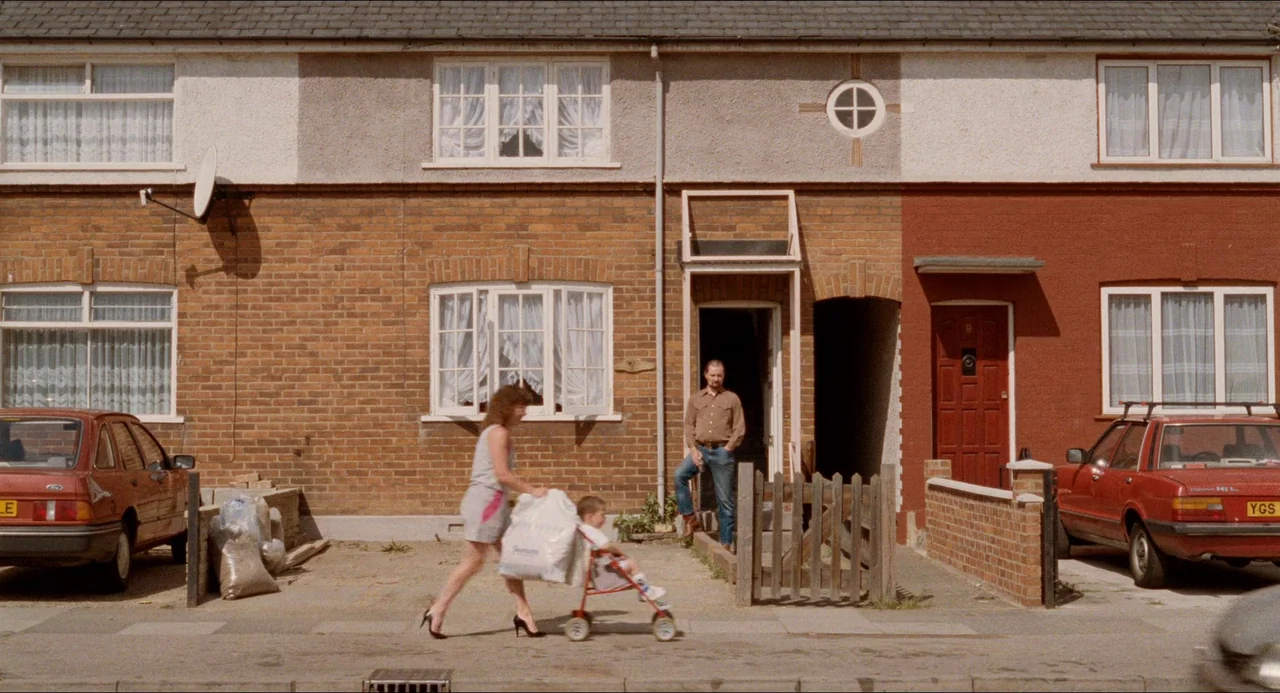 Life Is Sweet (1990) movie still: wide — A suburban row house street features brick exteriors and two parked red cars. A woman walks a stroller past…