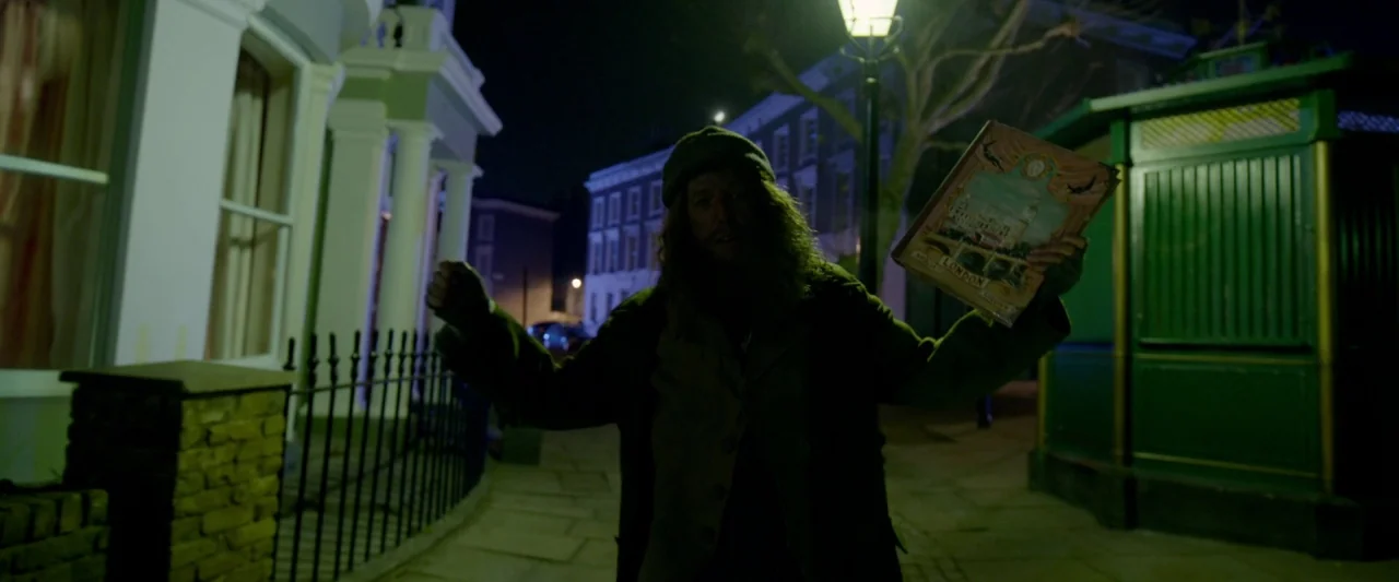 Paddington 2 (2017) movie still: medium — A dimly lit London street features Victorian-style residential buildings and a green kiosk. A bearded man…