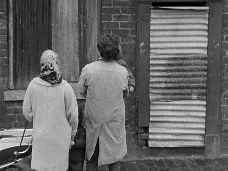 Cathy Come Home (1969) movie still: fullbody — An urban exterior features a brick wall with windows and a doorway boarded up. Two adults and a child…