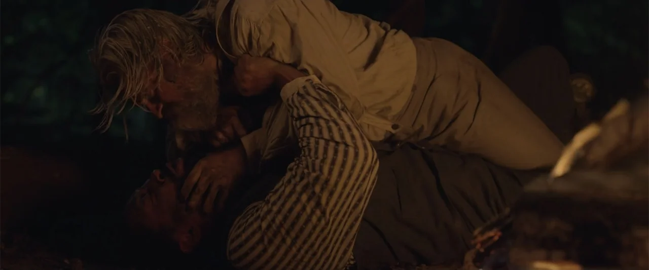 Ghosts of the Ozarks (2022) movie still: medium — A dark outdoor space at night illuminated by flickering light. Two men wrestle on the ground in a…
