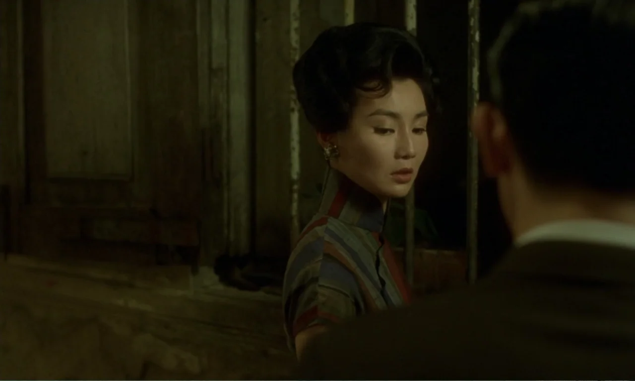 In the Mood for Love (2000) movie still: medium — A dimly lit indoor hallway features iron window bars and weathered wooden walls. A woman in a…