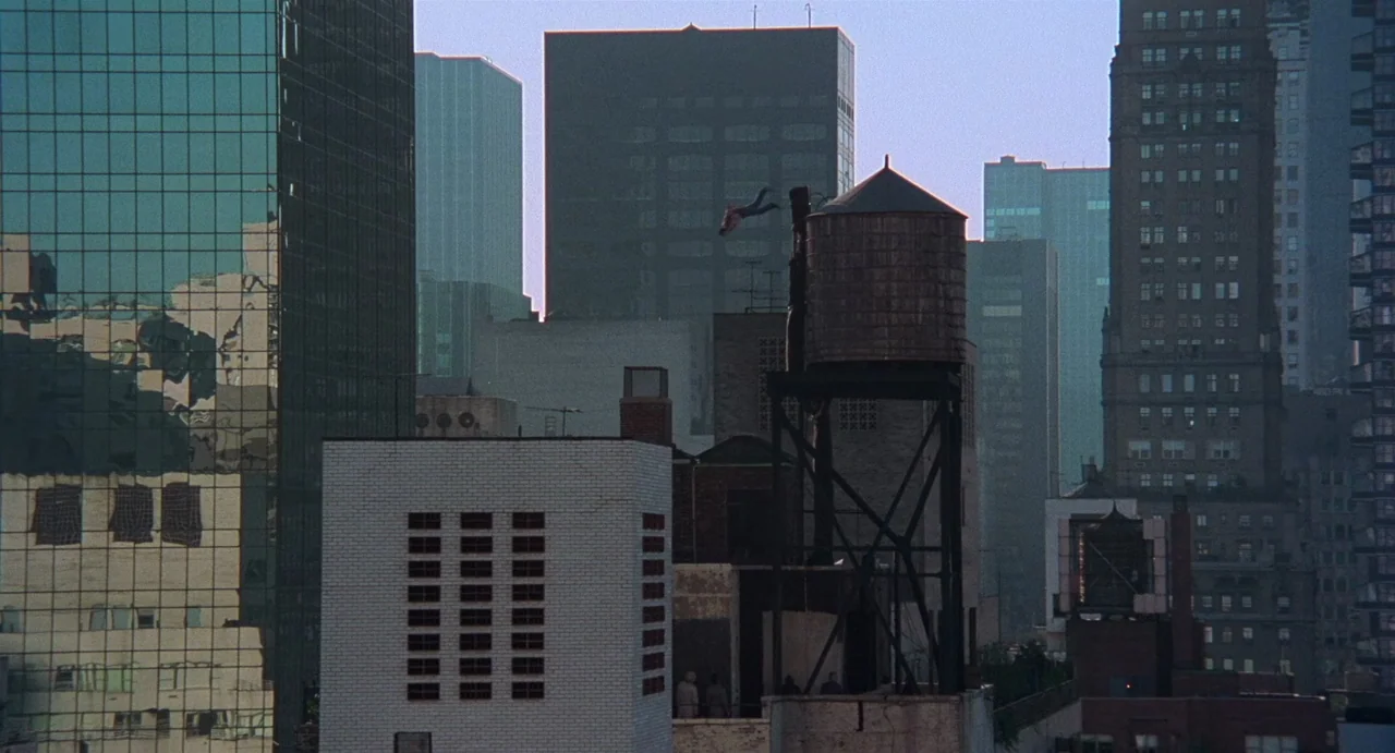 God Told Me To (1976) movie still: wide — A dense urban rooftop setting with several water towers and brick buildings. A costumed figure leaps…