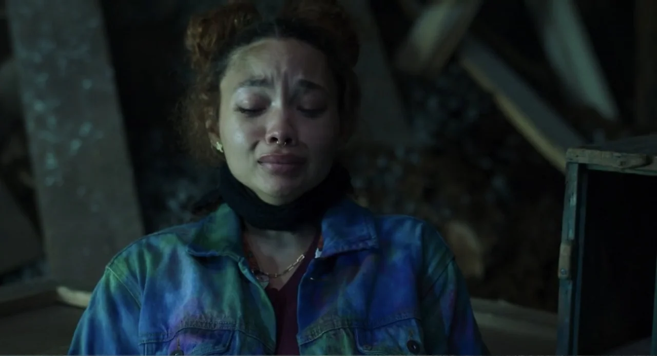 Salvation (2025) movie still: closeup — A dimly lit room with cluttered wooden structures in the dark background. A young woman cries while looking…