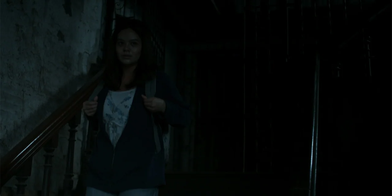 Nokturno (2024) movie still: medium — A dimly lit indoor staircase features dark wooden railings and weathered stone wall textures. A young woman…