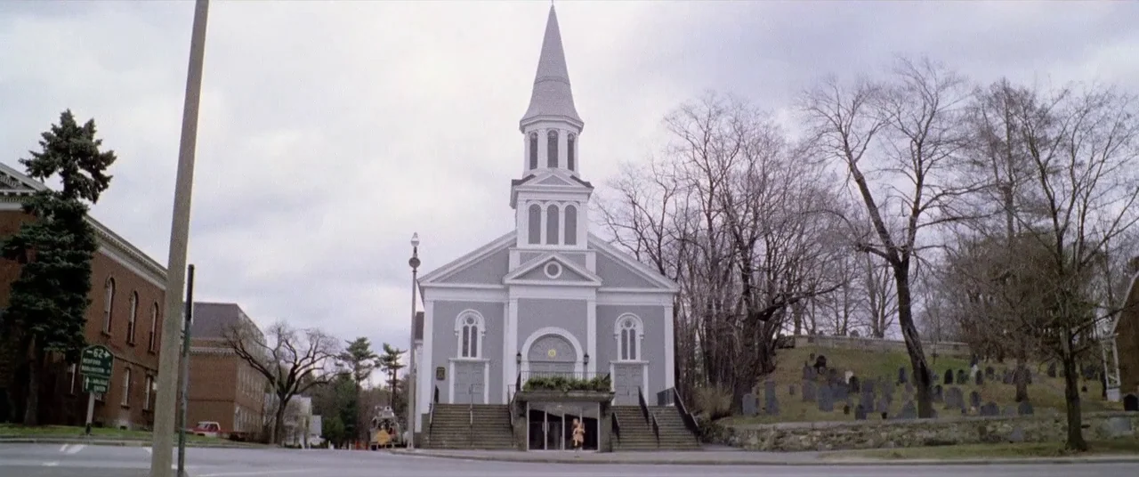 House By The Cemetery (1981) movie still: establishing — A light grey wooden church with a tall steeple stands near a cemetery. Bare trees and…