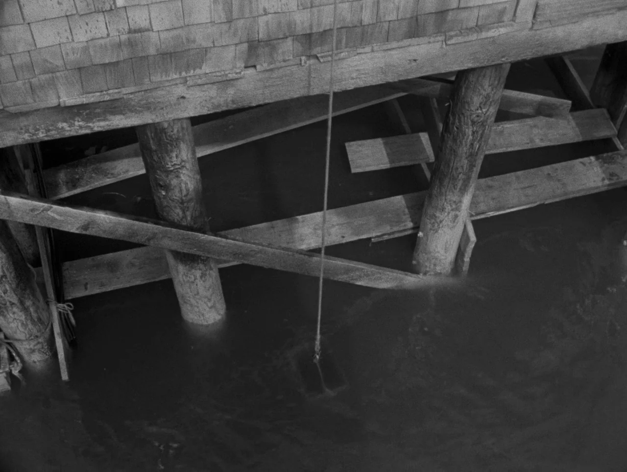 Pickup on South Street (1953) movie still: establishing — A wooden pier structure sits above calm, dark water with visible support pilings. A single…