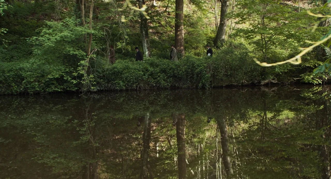 The Taste of Things (2023) movie still: wide — A quiet riverbank is bordered by a lush, green forest with dense undergrowth. Four men in early…