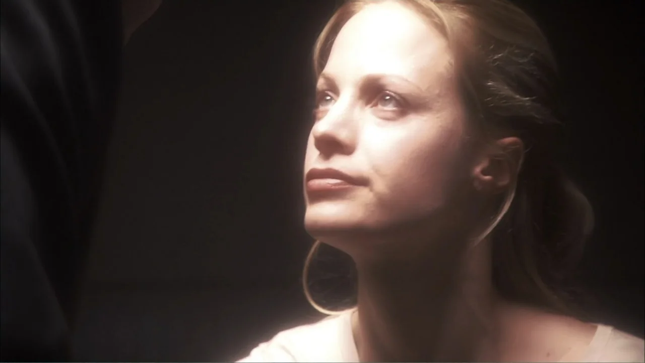 O código oculto (2025) movie still: closeup — A dark, unidentifiable interior space surrounds the subjects. A blonde woman looks upward at an unseen…