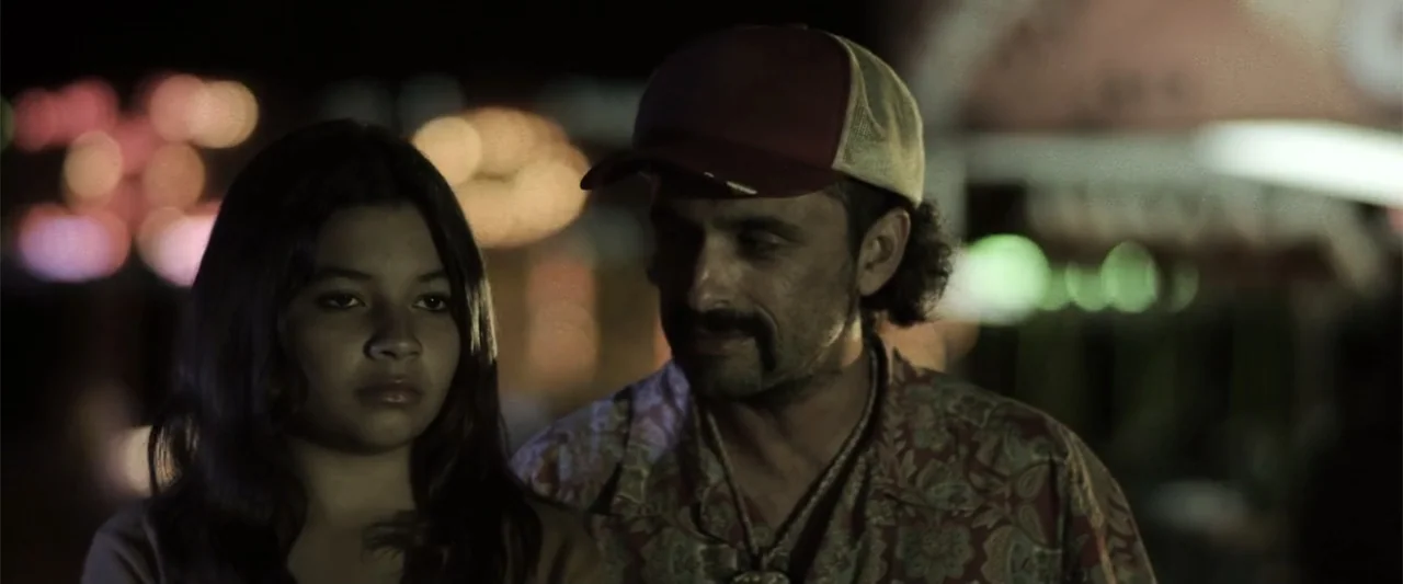 At Dawn (2013) movie still: medium — An outdoor fair at night with blurred light bokeh in the dark background. A man and a young girl stand side by…