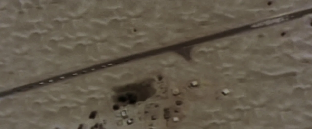 Syriana (2005) movie still: establishing — A vast expanse of sandy desert terrain is crossed by a straight paved road. Small clustered structures…