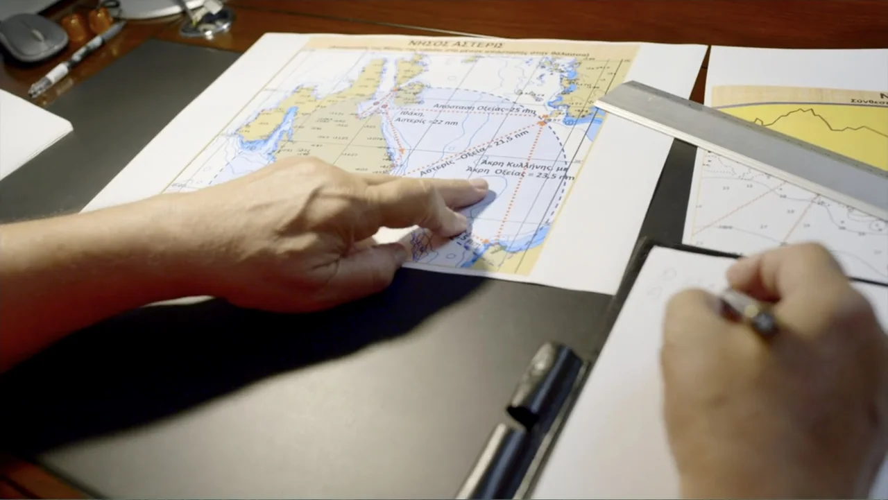 The Atlantis Puzzle (2024) movie still: closeup — An office desk features a nautical map, a metallic ruler, and a notepad. Hands are visible while…