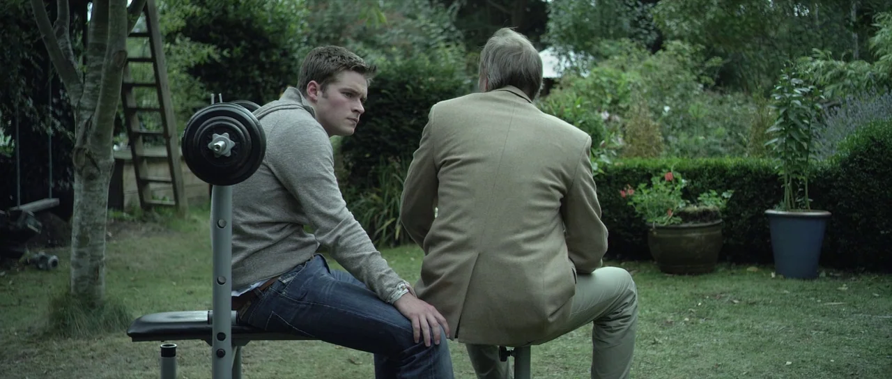 What Richard Did (2012) movie still: medium — A green residential lawn is filled with dense shrubs, trees, and potted plants. A young man and an…