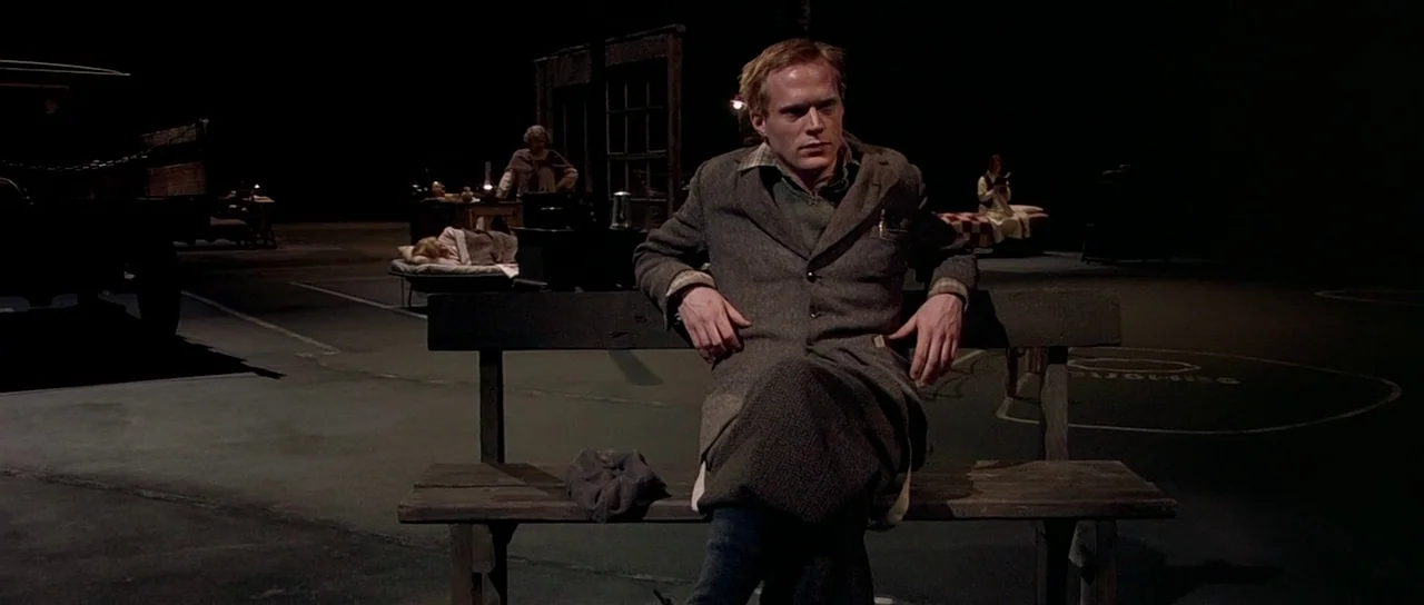 Dogville (2003) movie still: medium — A minimalist studio set depicts a dark open space with scattered furniture items. A man sits prominently on a…