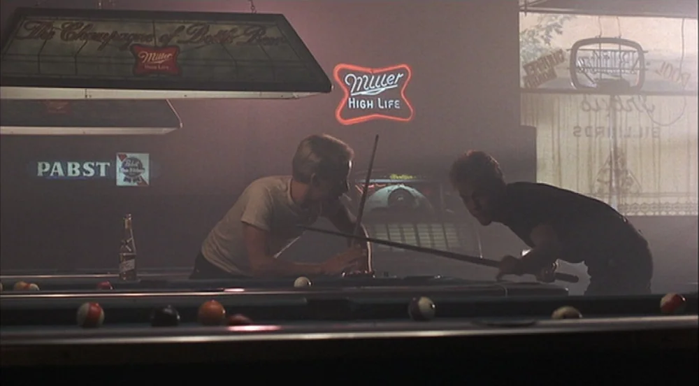 Stand By Me (1986) movie still: A quiet, atmospheric moment in a dimly lit pool hall where two young men lean into a game of billiards. The air is…