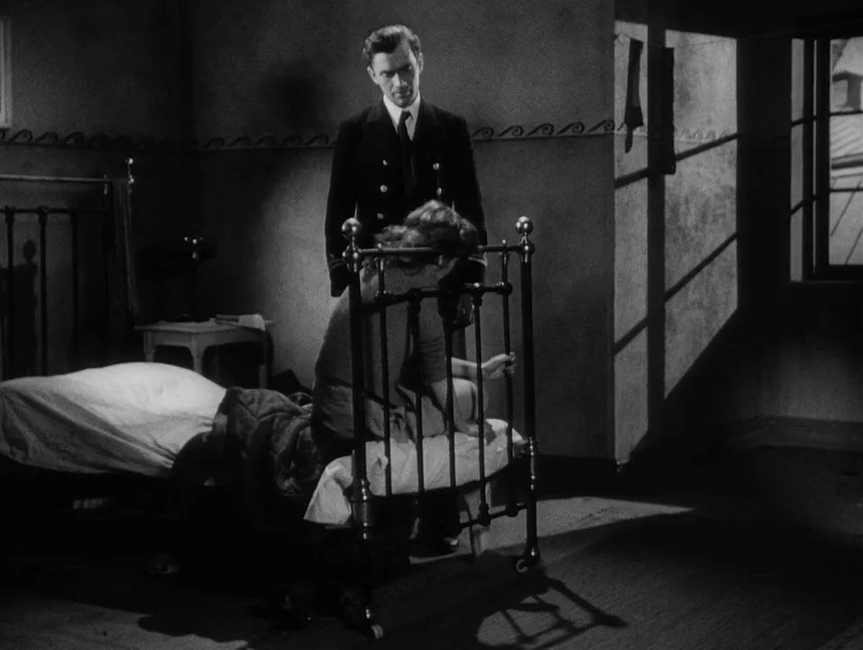 A Ship Bound for India (1947) movie still: A tense, dramatic moment unfolding in a spartan, shadow-heavy room. The stark, high-contrast lighting…