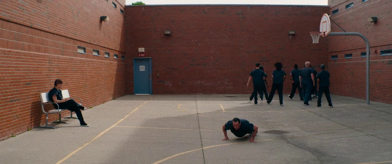 Dinner in America (2020) movie still: A stark, wide-angle view of a desolate prison courtyard under harsh daylight. The composition emphasizes the…