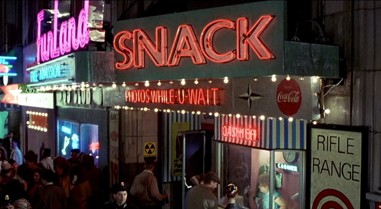 Dogfight (1991) movie still: wide — A dense urban arcade storefront at night features glowing red and blue neon signs. A crowd of people moves along…