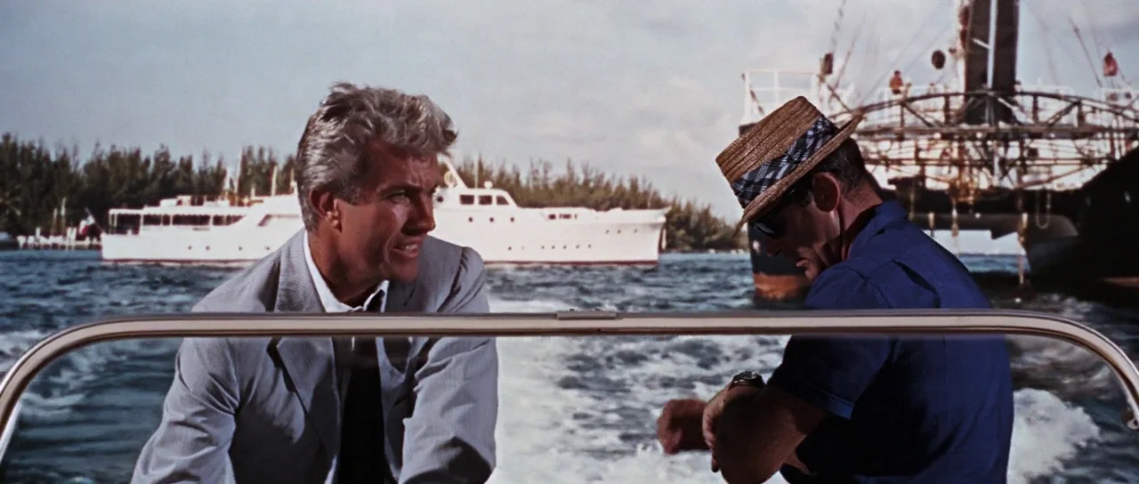 Thunderball (1965) movie still: medium — Two men ride on a motorboat on open water on a sunny day. A large white yacht and a construction platform…