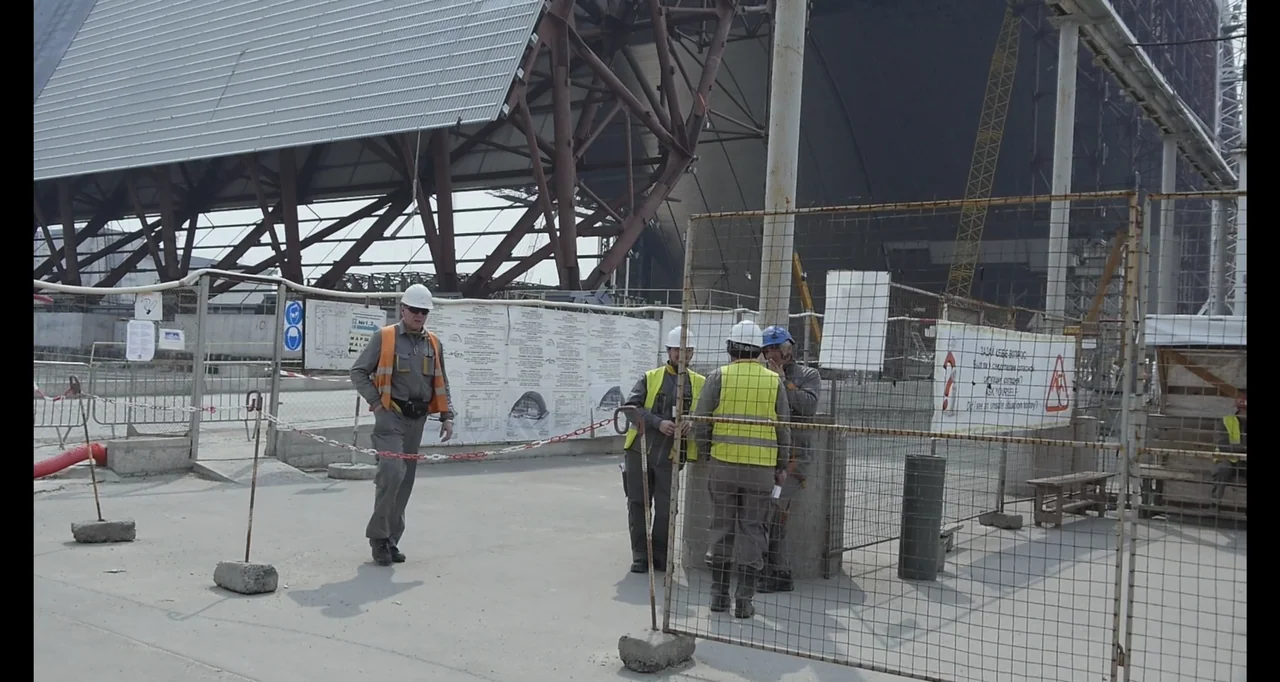 Chernobyl's Café (2016) movie still: wide — A vast industrial construction site features large metal framework structures and temporary chain-link…