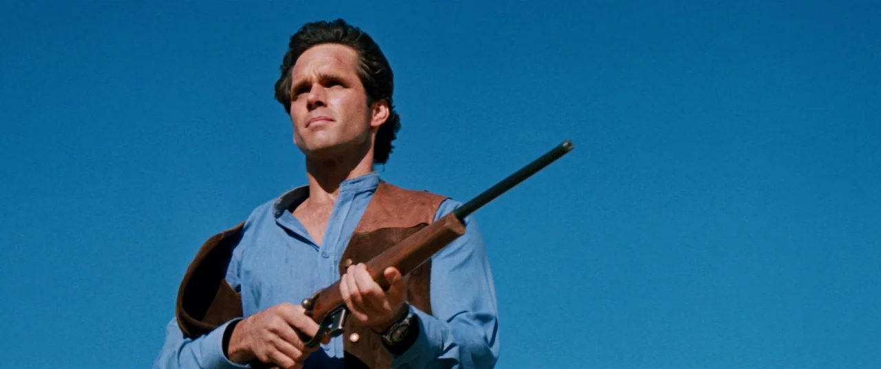 Razorback (1984) movie still: medium — A man stands outdoors under a clear deep blue sky. He holds a wooden hunting rifle across his chest while…