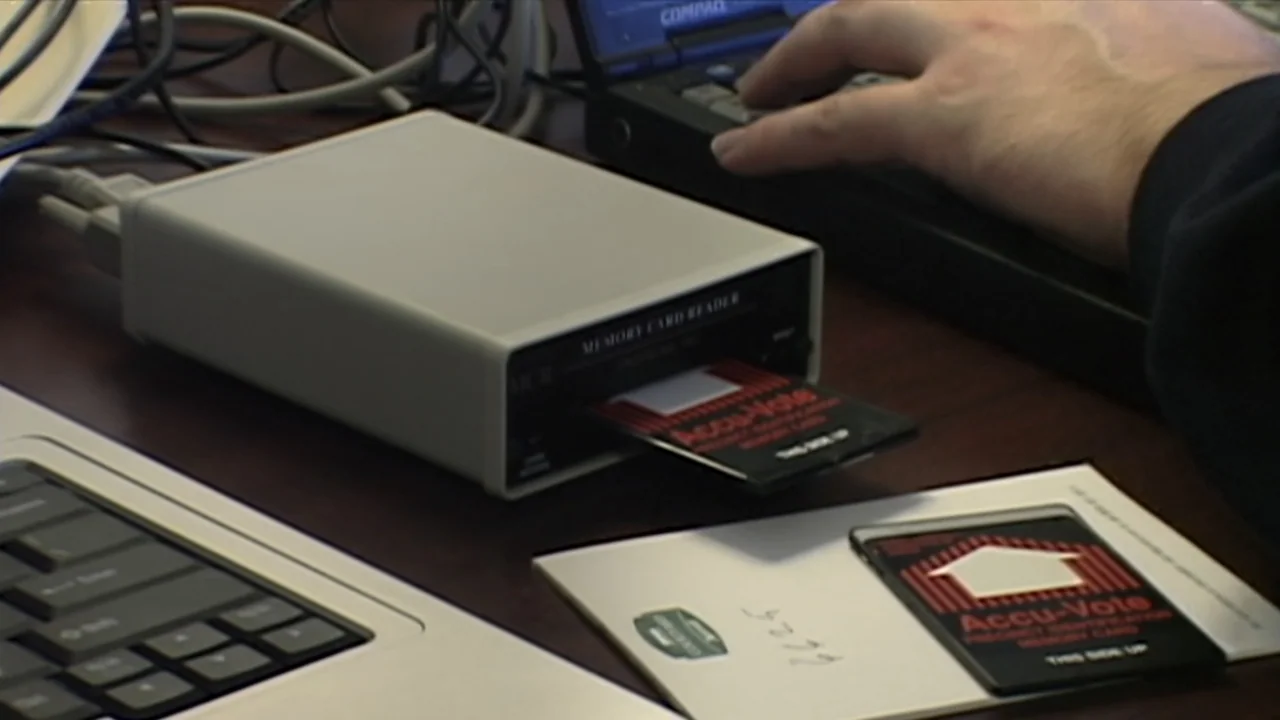Kill Chain: The Cyber War on America's Elections (2020) movie still: medium — A dark wooden desk holds a laptop and a gray memory card reader. A…