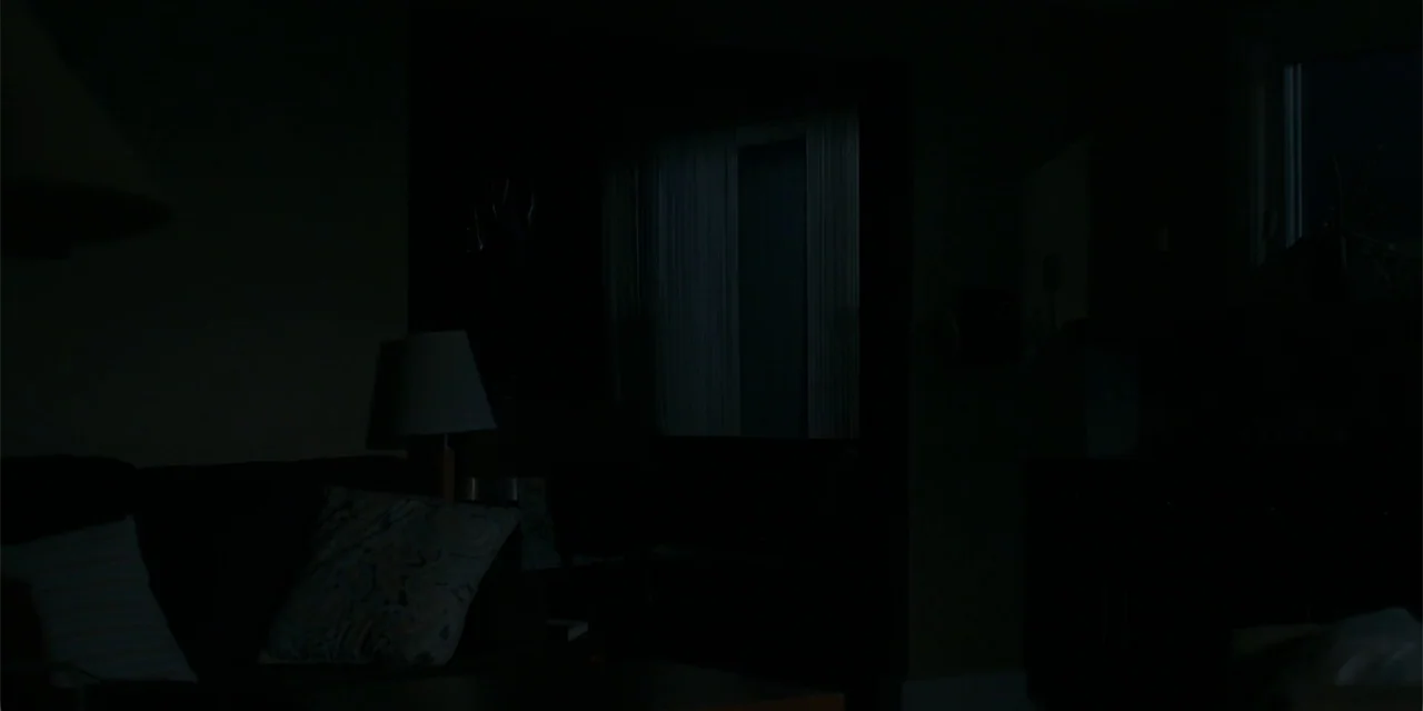Demonic (2015) movie still: establishing — A dimly lit room interior features a sofa and a lamp at night. The space is mostly obscured by deep…