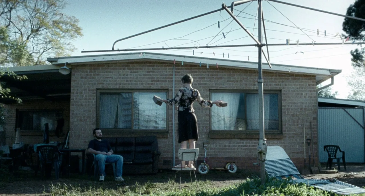 Snowtown (2011) movie still: wide — A suburban brick house stands behind a neglected backyard featuring a rotary clothesline. A woman balances atop…