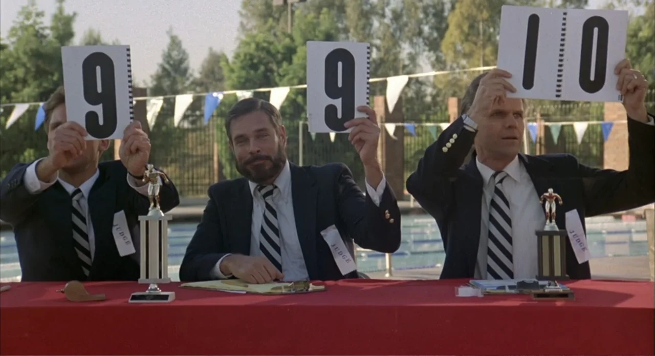 Back to School (1986) movie still: medium — Three men in formal attire sit at a red table before a swimming pool. The men hold up white scorecards…