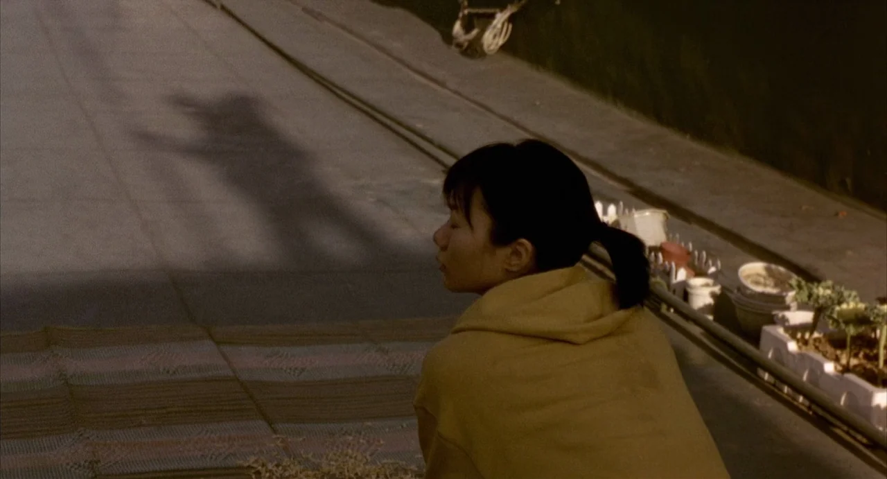 Barking Dogs Never Bite (2000) movie still: medium — A rooftop surface covered with a woven mat features a young woman sitting down. A long shadow…