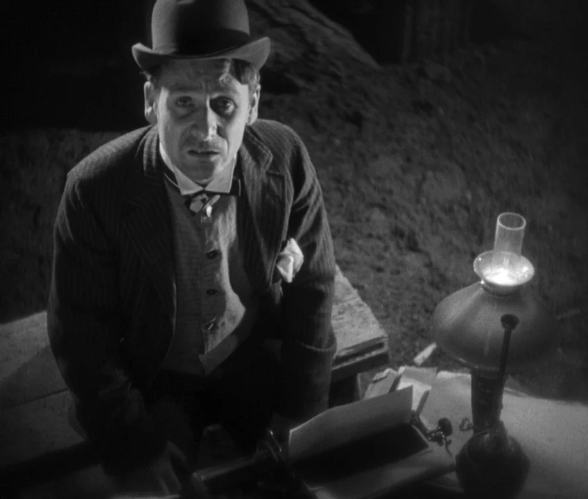 The Threepenny Opera (1931) movie still: medium — A dark, sparsely lit workspace features a wooden desk with a vintage typewriter. A man wearing a…