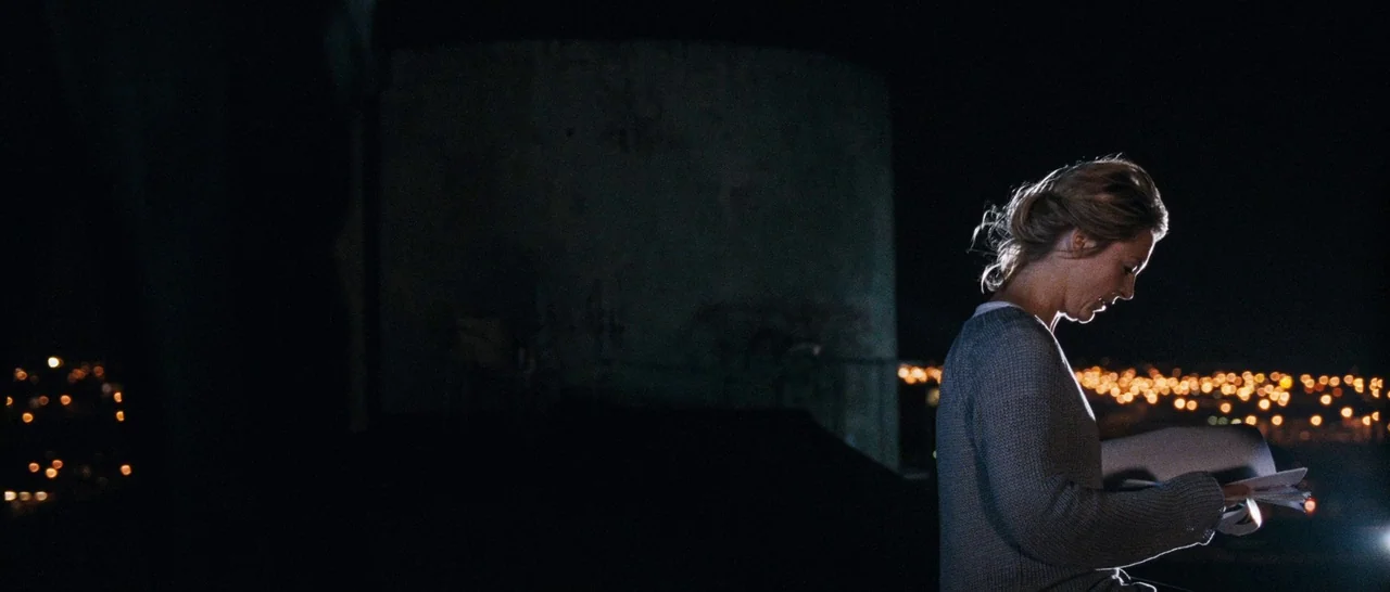 Sound of Noise (2010) movie still: medium — A woman stands on a dark, desolate rooftop at night overlooking distant city lights. She reads a stack…